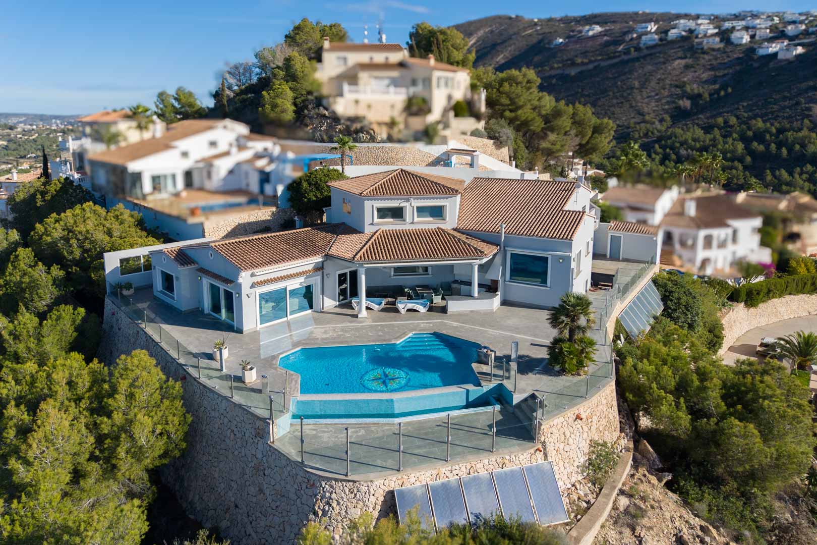 Vista aérea de la villa con piscina azul, terraza cubierta y tejado de tejas, rodeada de jardines y muros de piedra.