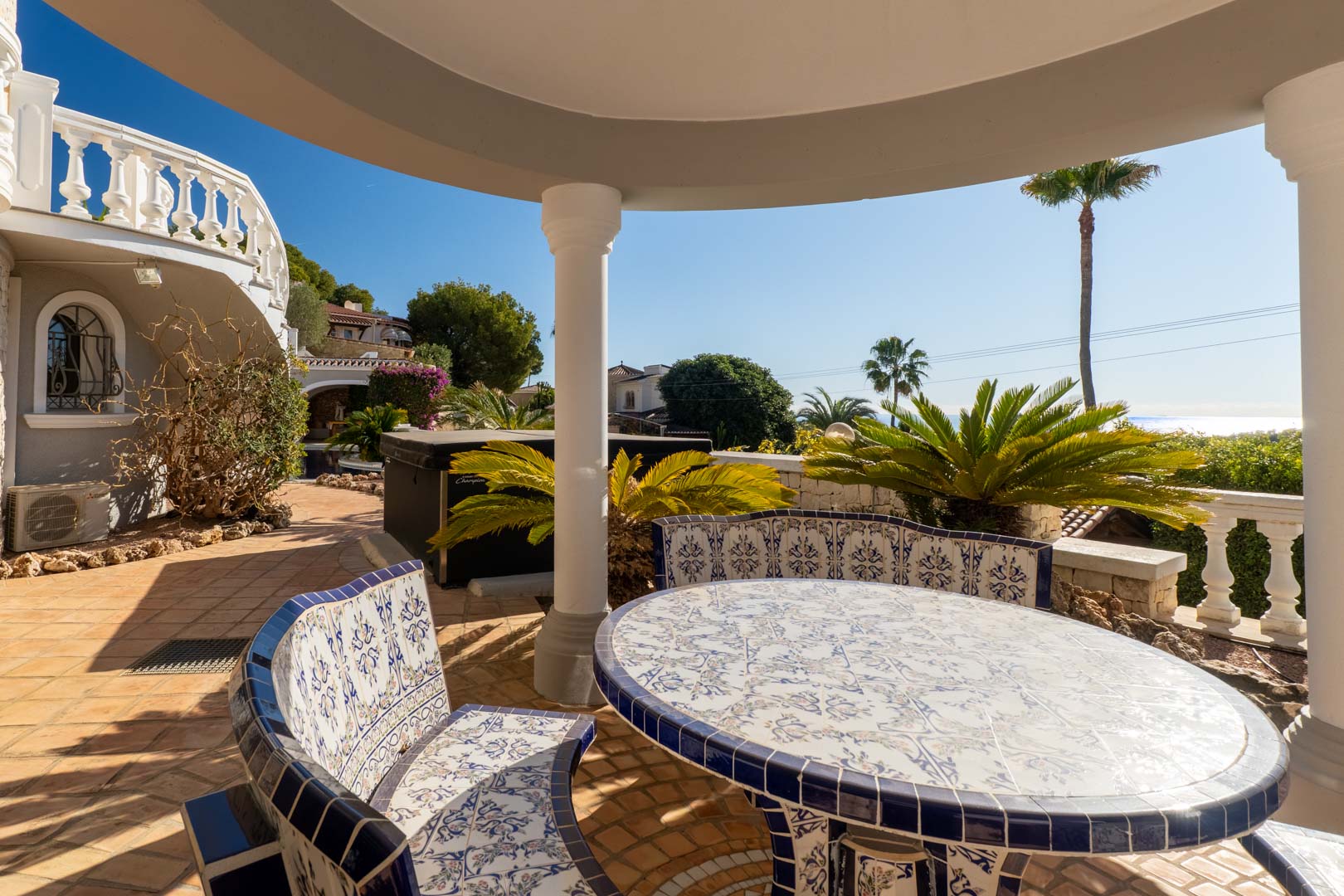 Área de comedor exterior bajo porche con mesa y bancos curvos decorados con azulejos tradicionales en azul y blanco, con vistas al jardín mediterráneo y al mar.