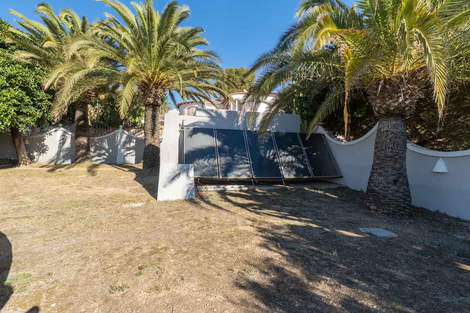 Área de jardín con instalación de paneles solares térmicos, rodeada de palmeras maduras y muros perimetrales blancos bajo luz natural directa.