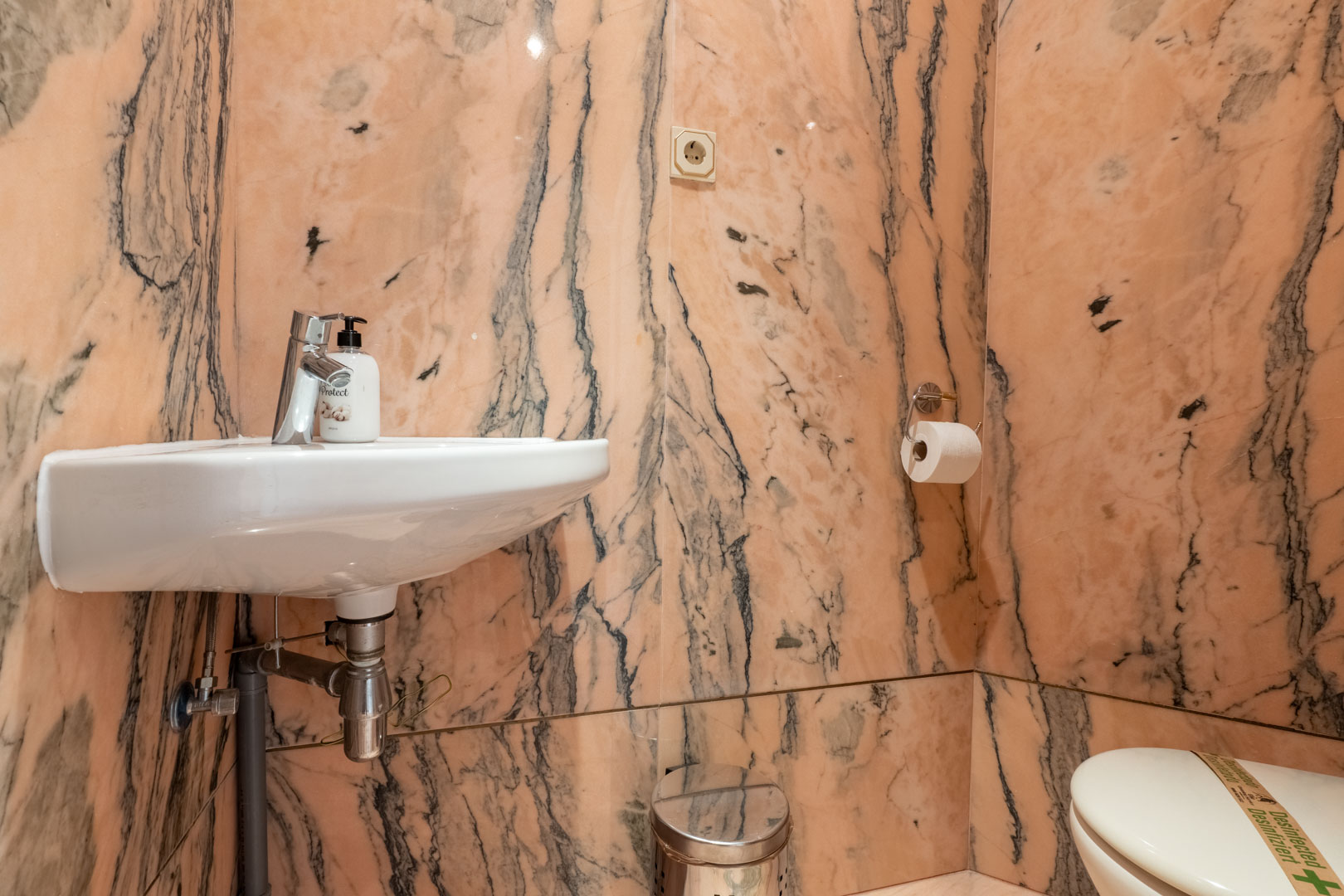 Aseo de invitados revestido íntegramente en mármol rosa con vetas oscuras, equipado con un lavabo suspendido blanco y grifería cromada.