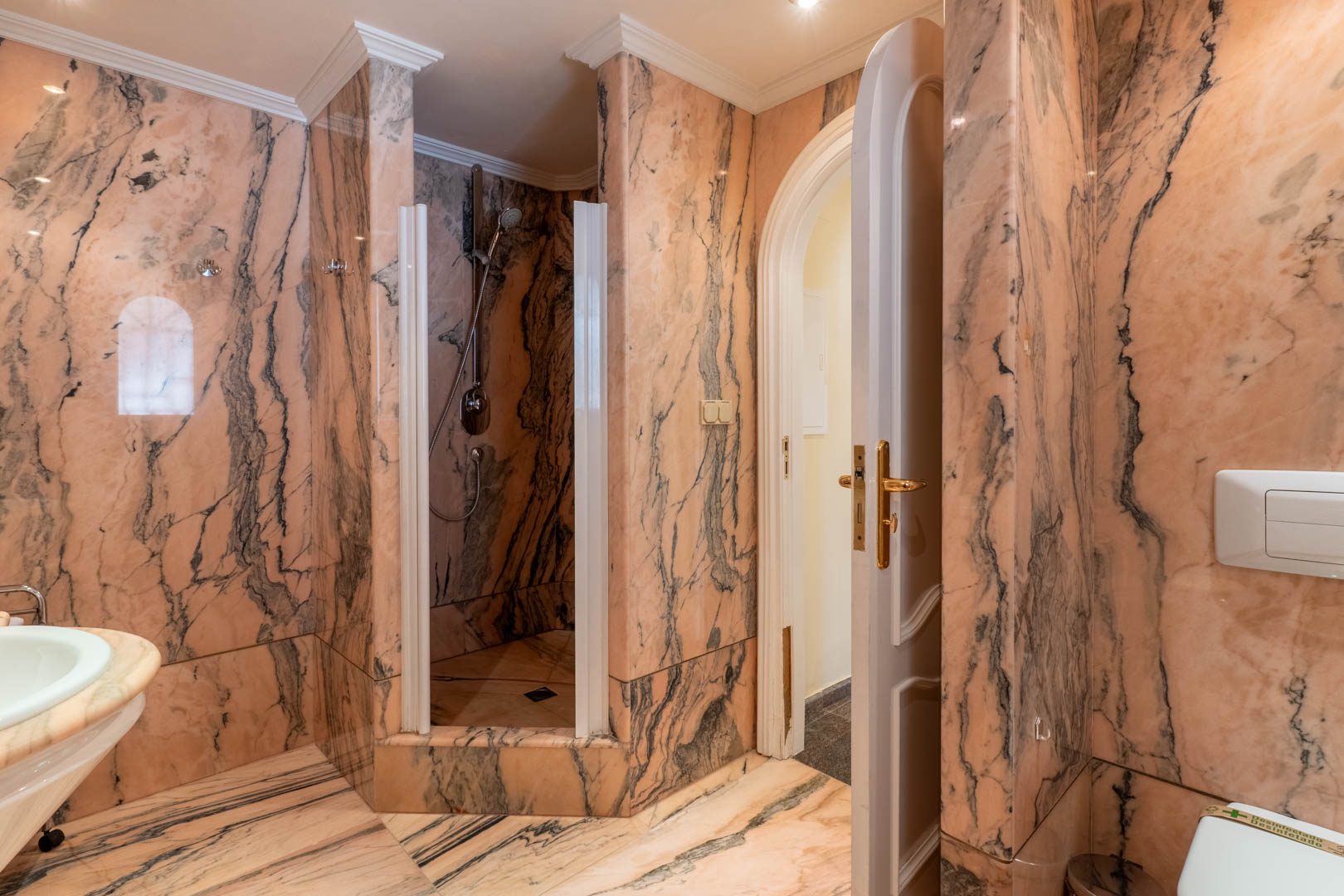 Baño revestido íntegramente en mármol rosa con vetas oscuras, que incluye una ducha de obra, molduras clásicas y una puerta de paso en arco con herrajes dorados.