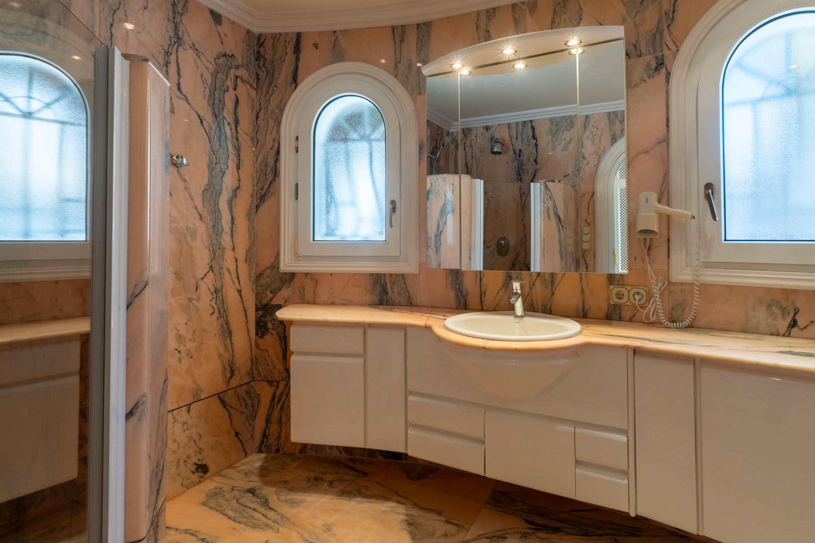 Baño revestido íntegramente en mármol rosado con vetas grises, equipado con un mueble de lavabo blanco y ventanas de arco con cristal translúcido.