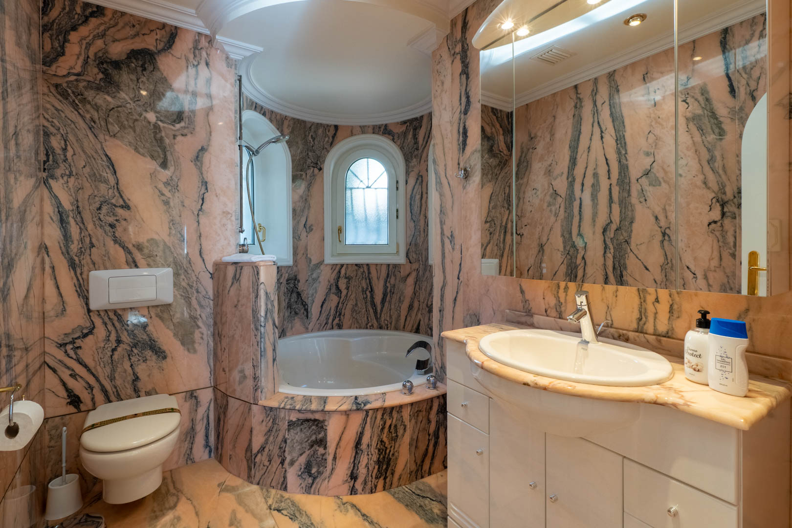 Baño revestido íntegramente en mármol veteado en tonos salmón y gris, con bañera encastrada en un nicho curvo, ventanas de arco y mueble de lavabo blanco con encimera de piedra.