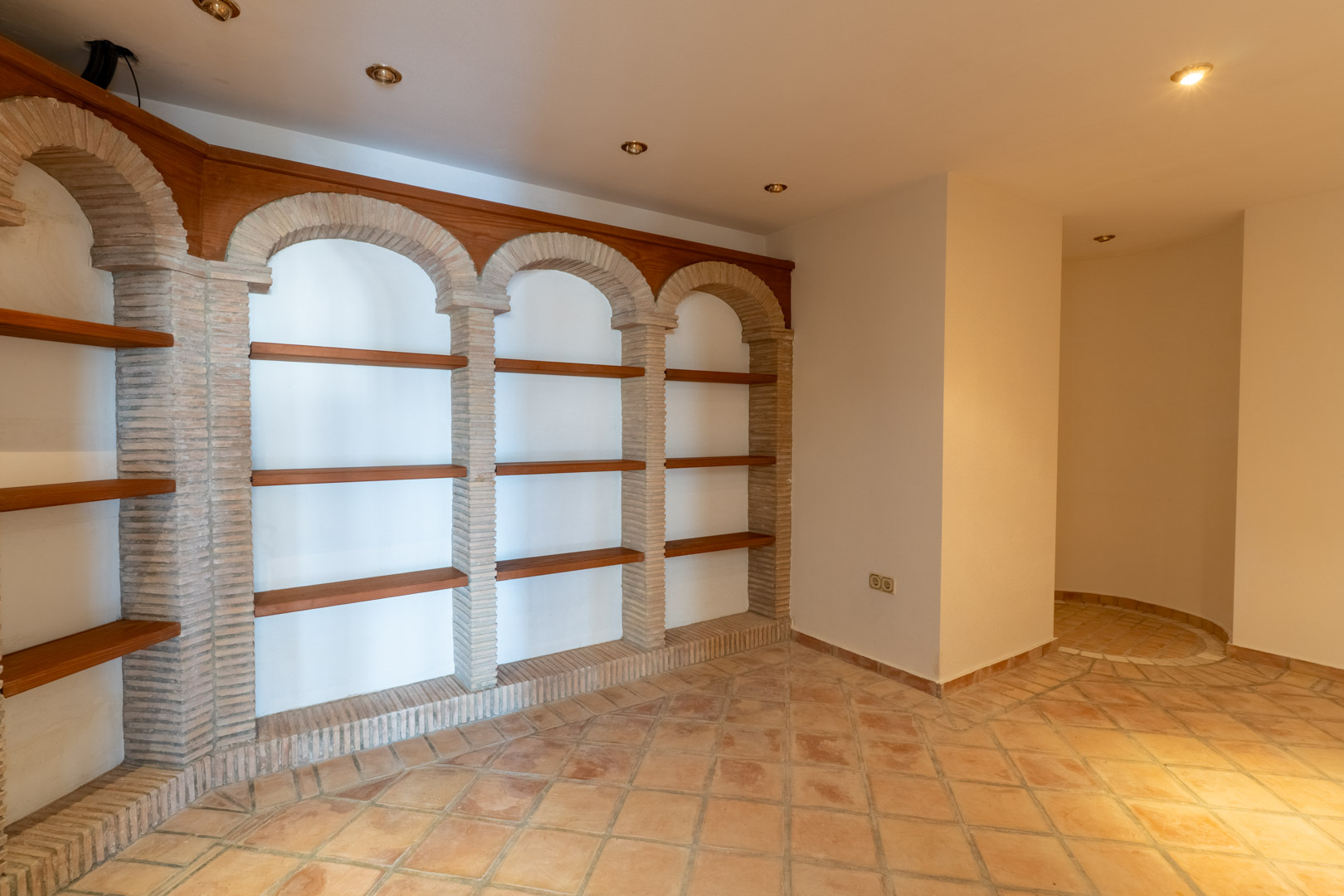 Bodega con estanterías de madera integradas en arcos de ladrillo visto, suelos de terracota y luminarias empotradas en el techo.