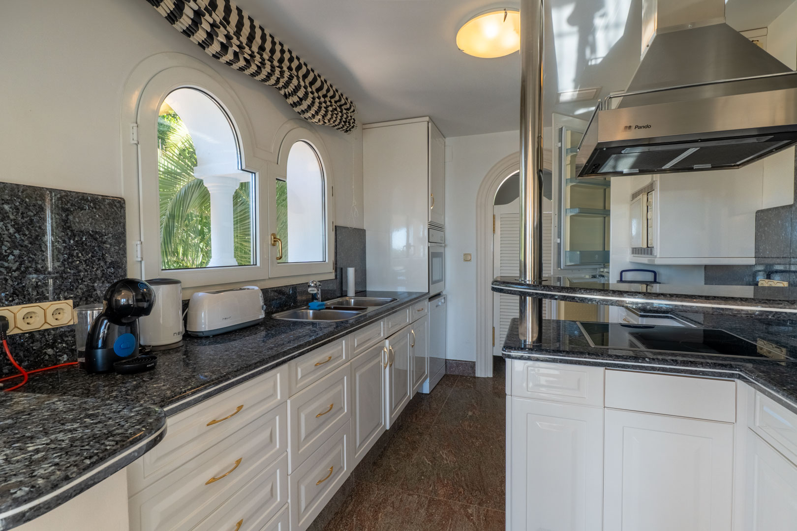 Cocina independiente con encimeras de granito oscuro, mobiliario blanco y ventanas de arco con vistas al jardín. Incluye campana extractora Pando y electrodomésticos integrados.