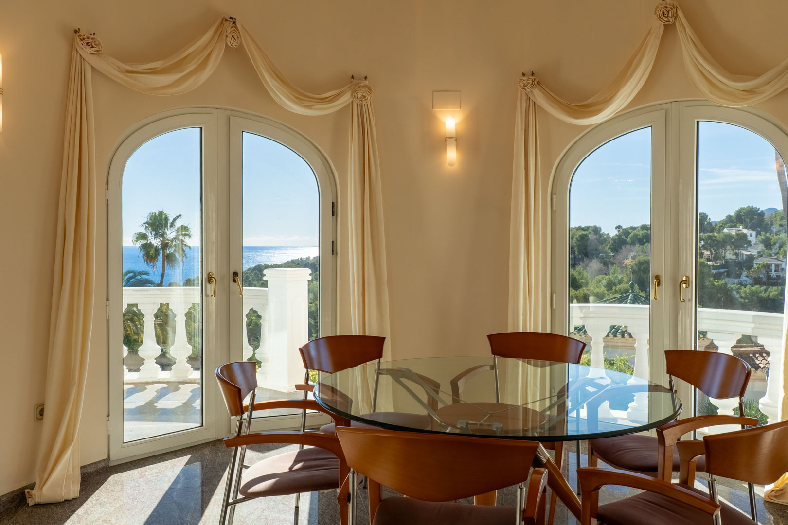 Comedor con mesa de cristal y sillas de madera, rodeado de ventanales en arco que ofrecen vistas panorámicas al mar y acceso directo a la terraza con balaustrada.
