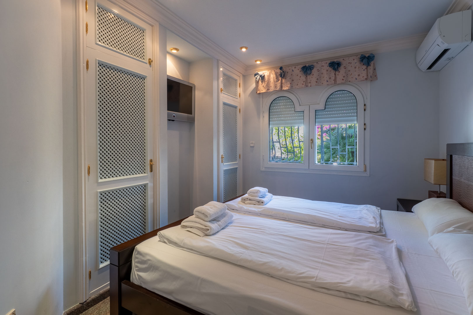 Dormitorio con armarios empotrados de rejilla blanca, televisor integrado en pared y ventana de doble arco con vistas al jardín.