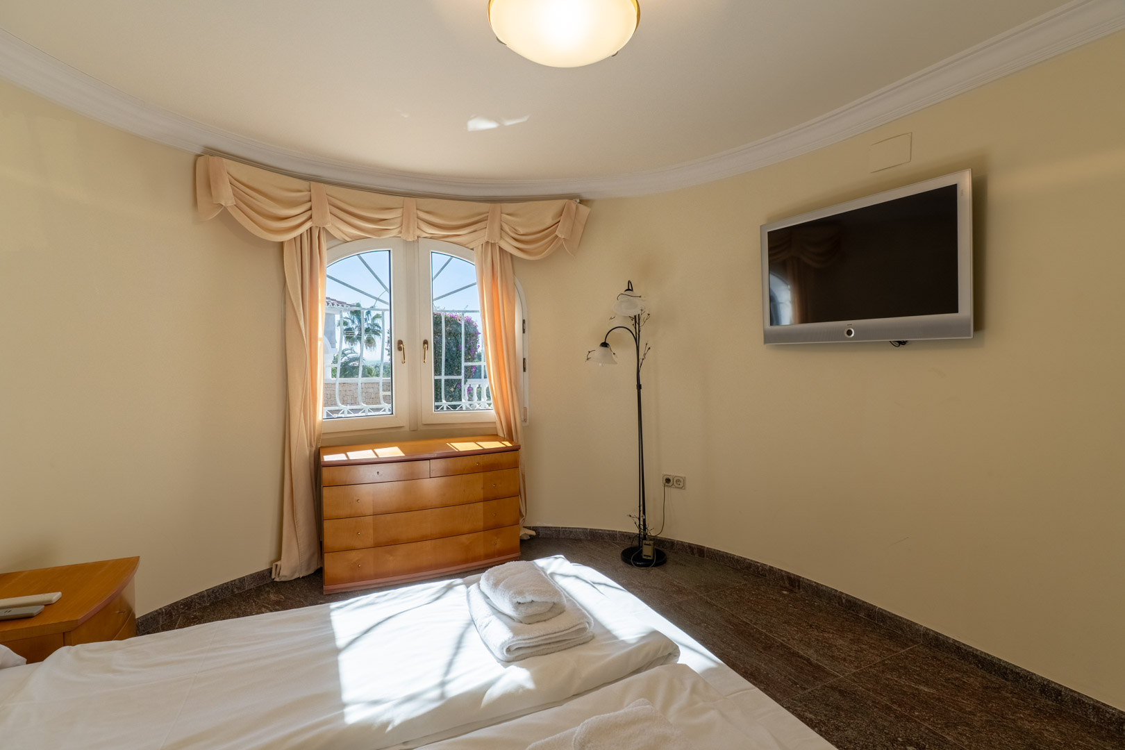Dormitorio con pared curva, suelo de granito oscuro y ventana de arco con cortinas clásicas. Incluye cómoda de madera, TV de pared y abundante luz natural.