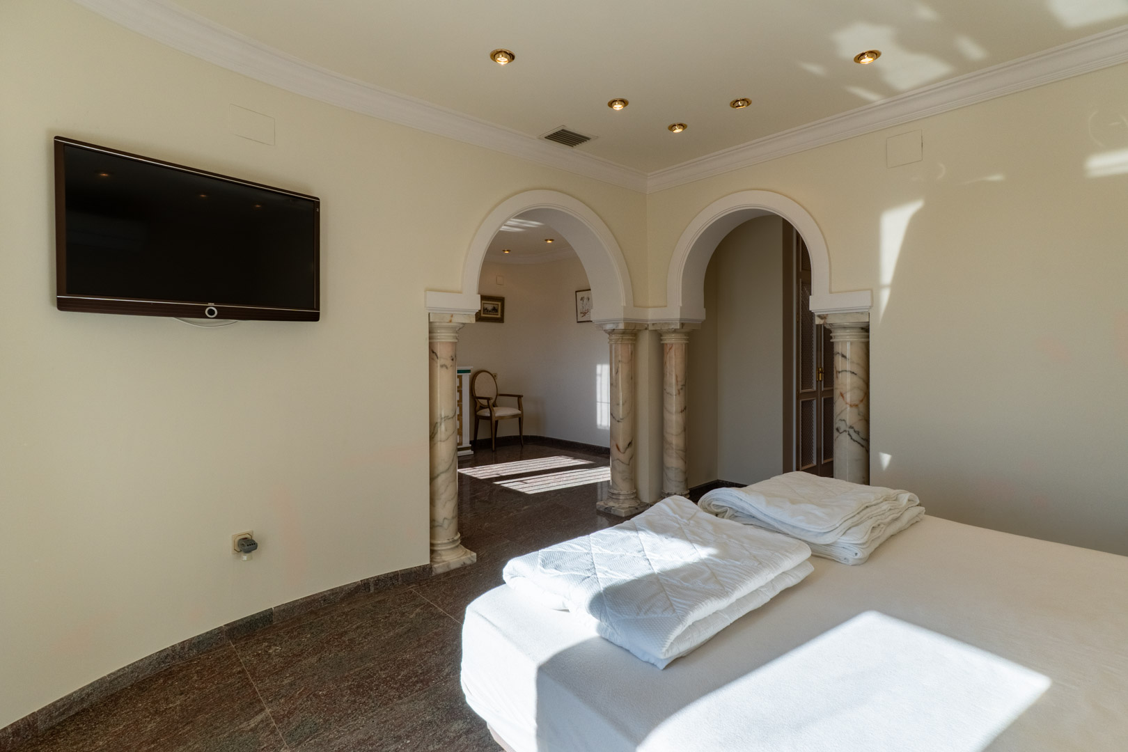 Dormitorio principal con arcos de estilo clásico sobre columnas de mármol, suelos de piedra pulida y acceso a una estancia contigua bajo luz natural.