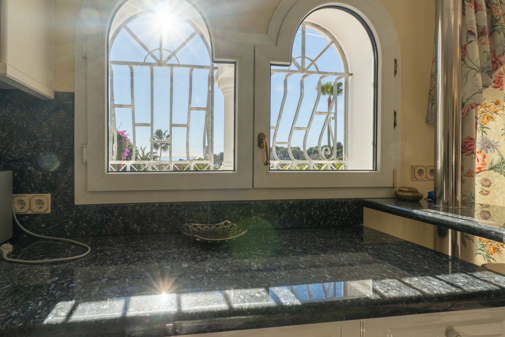 Encimera de granito oscuro bajo ventanas de arco con rejas decorativas, que ofrecen vistas al jardín mediterráneo y abundante luz natural.
