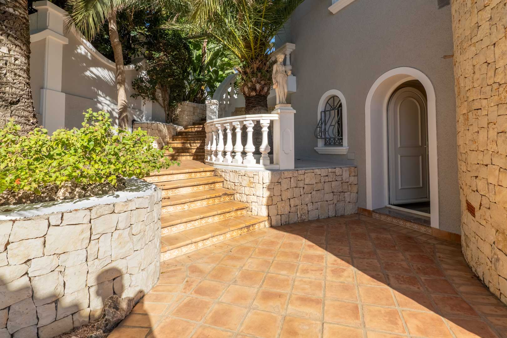 Entrada exterior con suelo de terracota, muros de piedra seca y una escalera con balaustrada. La arquitectura destaca por sus arcos y la frondosa vegetación mediterránea.