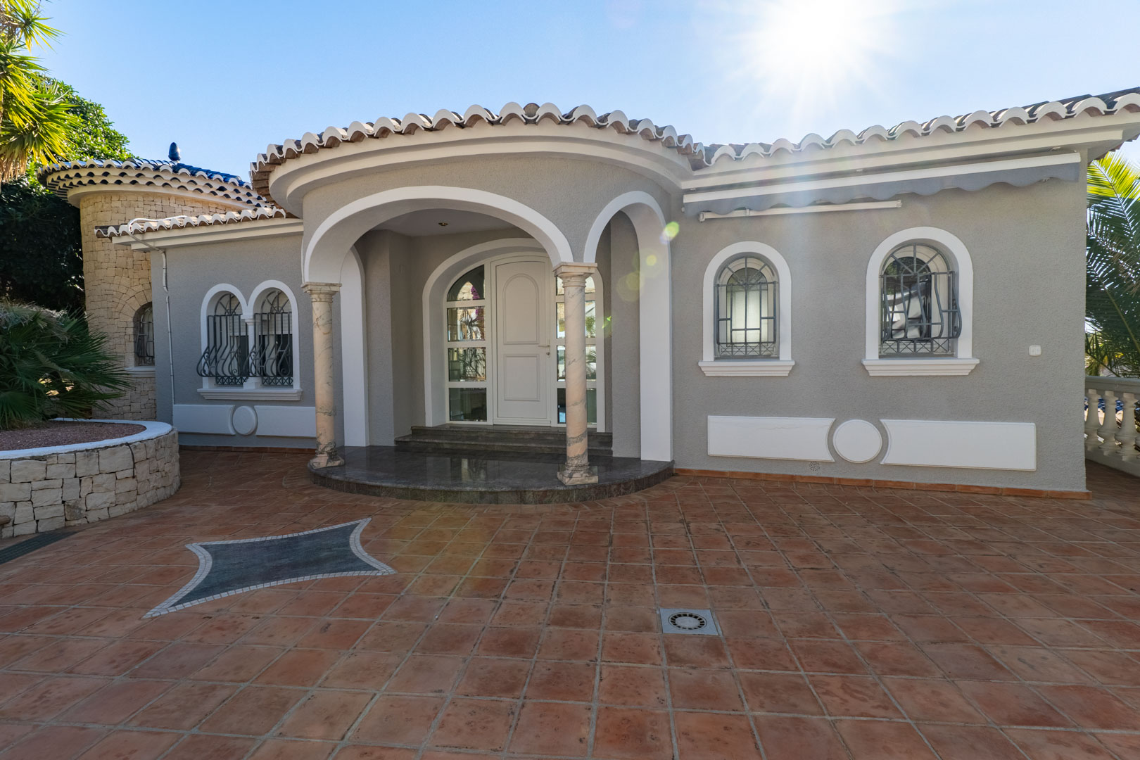 Entrada principal con porche de arcos sobre columnas de mármol, suelo de terracota con mosaico decorativo y fachada gris con molduras blancas y ventanas de forja.