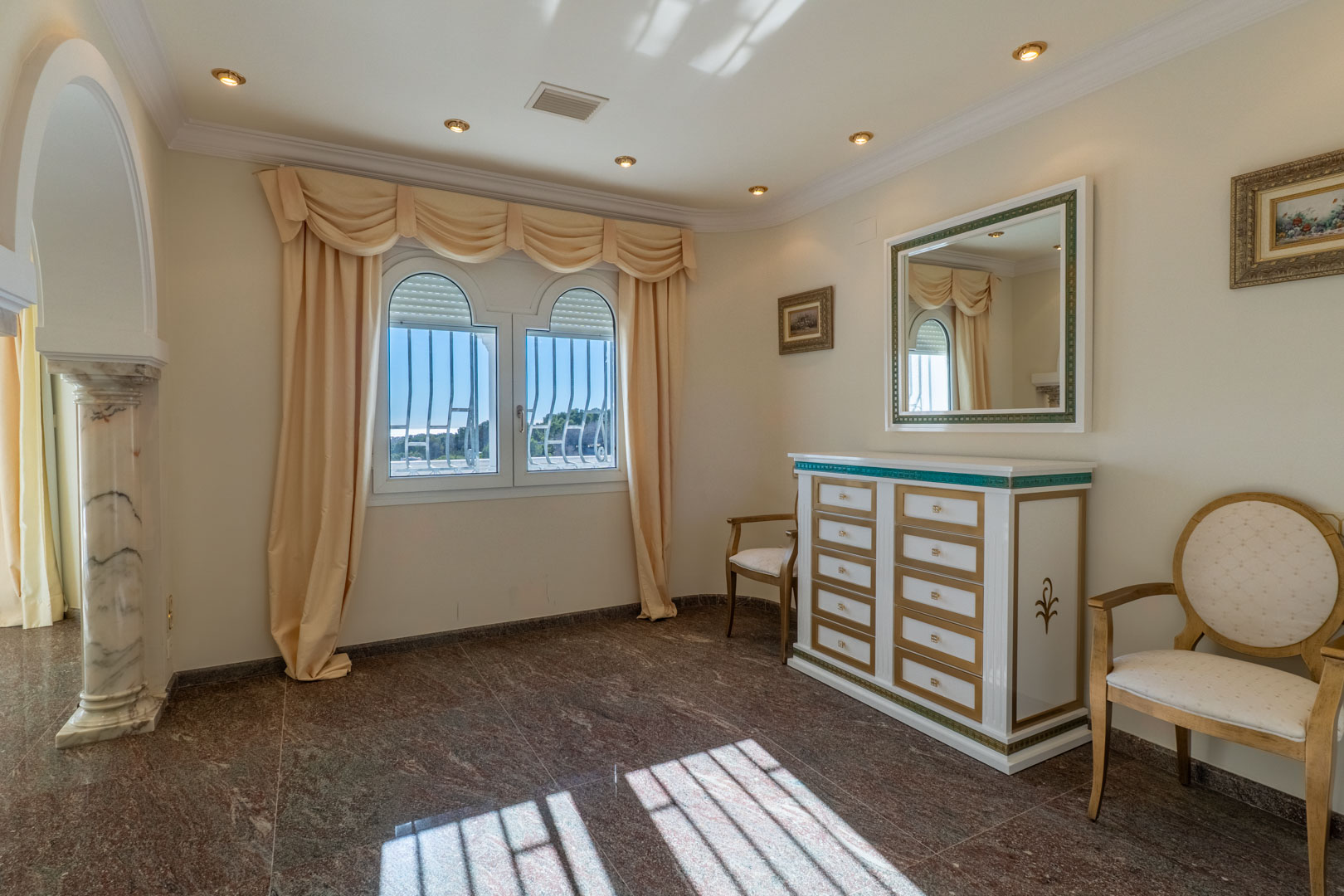 Estancia elegante con suelos de granito, cómoda de diseño clásico y una columna de mármol que enmarca el acceso junto a ventanales en arco.