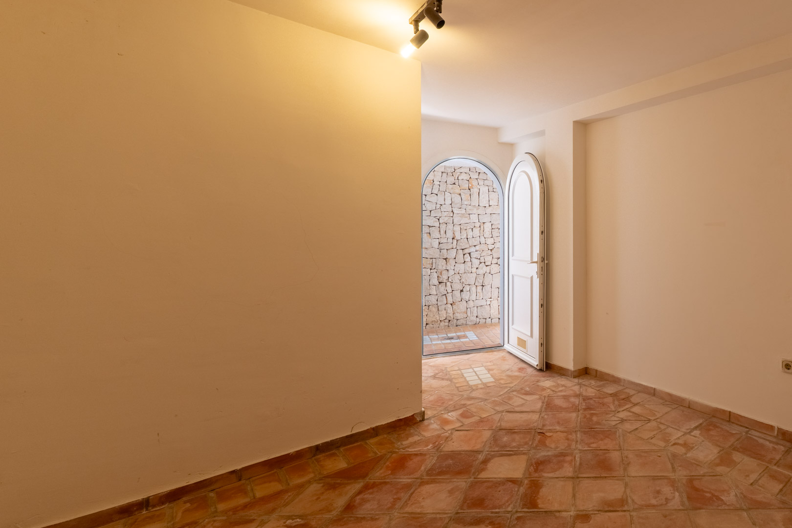 Habitación funcional con suelos de terracota y paredes blancas, que cuenta con una puerta de arco abierta hacia un patio con muro de piedra natural.