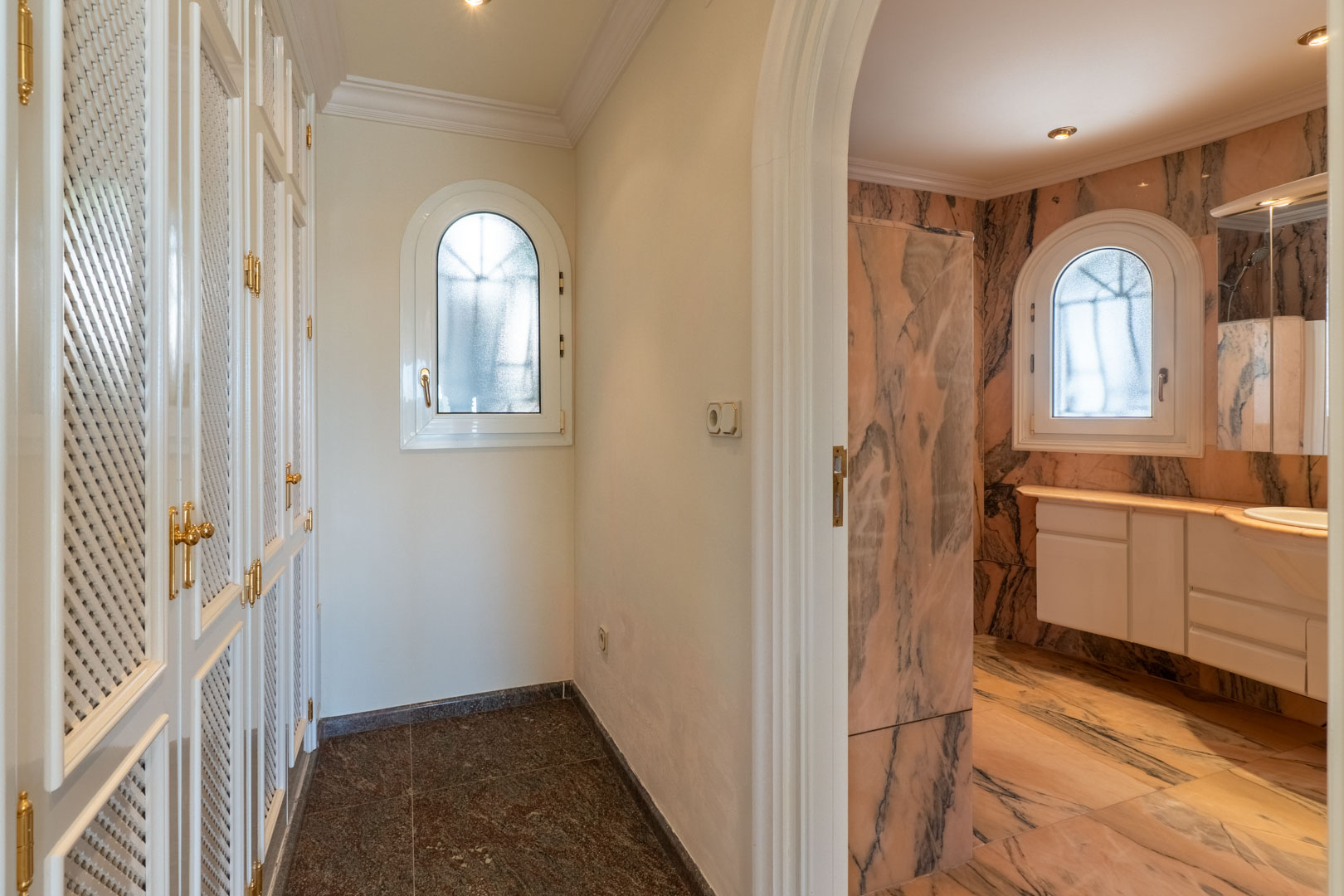 Pasillo con armarios empotrados de celosía blanca y herrajes dorados que da paso a un baño revestido íntegramente en mármol rosado con ventanas de arco.