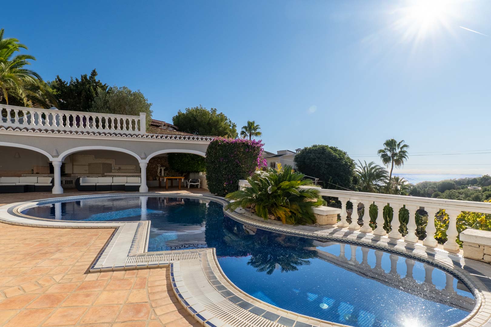 Piscina de diseño irregular con azulejos azules, rodeada de una terraza de barro y un porche con arcos blancos y vistas panorámicas al mar Mediterráneo.