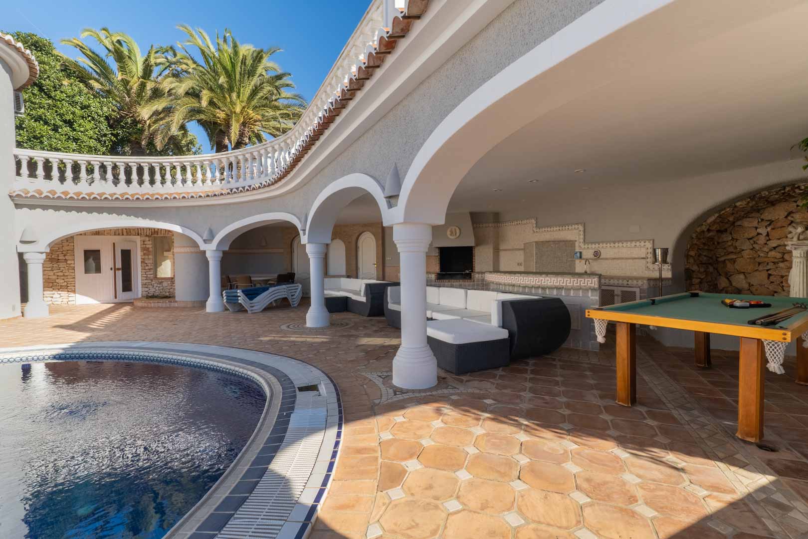 Terraza cubierta con arcos junto a la piscina, equipada con zona de estar, mesa de billar y cocina de verano integrada con acabados cerámicos.