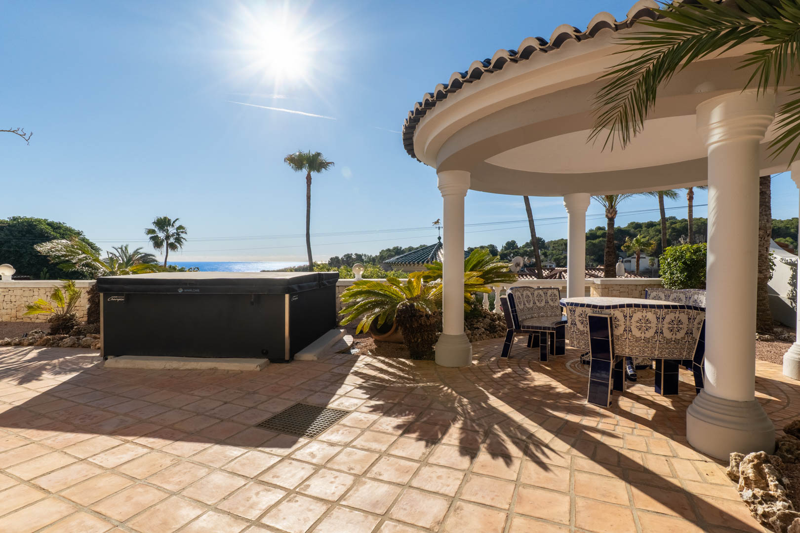 Terraza exterior con suelos de terracota, jacuzzi cubierto y un porche circular con columnas que resguarda un conjunto de mesa y bancos de cerámica tradicional frente al mar.