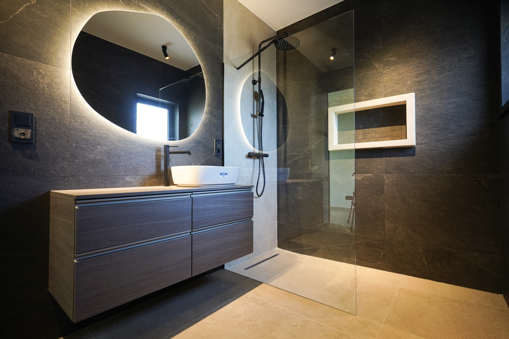 Baño de diseño con paredes en piedra oscura, lavabo de superficie y espejo orgánico retroiluminado. Dispone de ducha a ras de suelo con grifería negra y mampara de cristal.