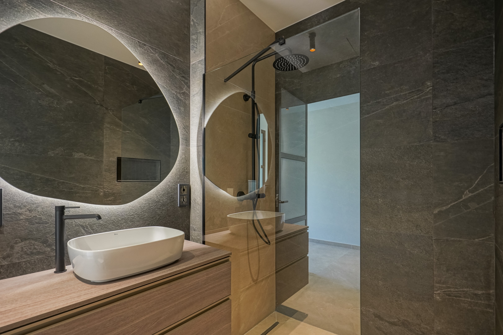 Baño moderno con azulejos de efecto piedra oscura, lavabo sobre encimera de madera y espejo circular retroiluminado junto a una ducha italiana.