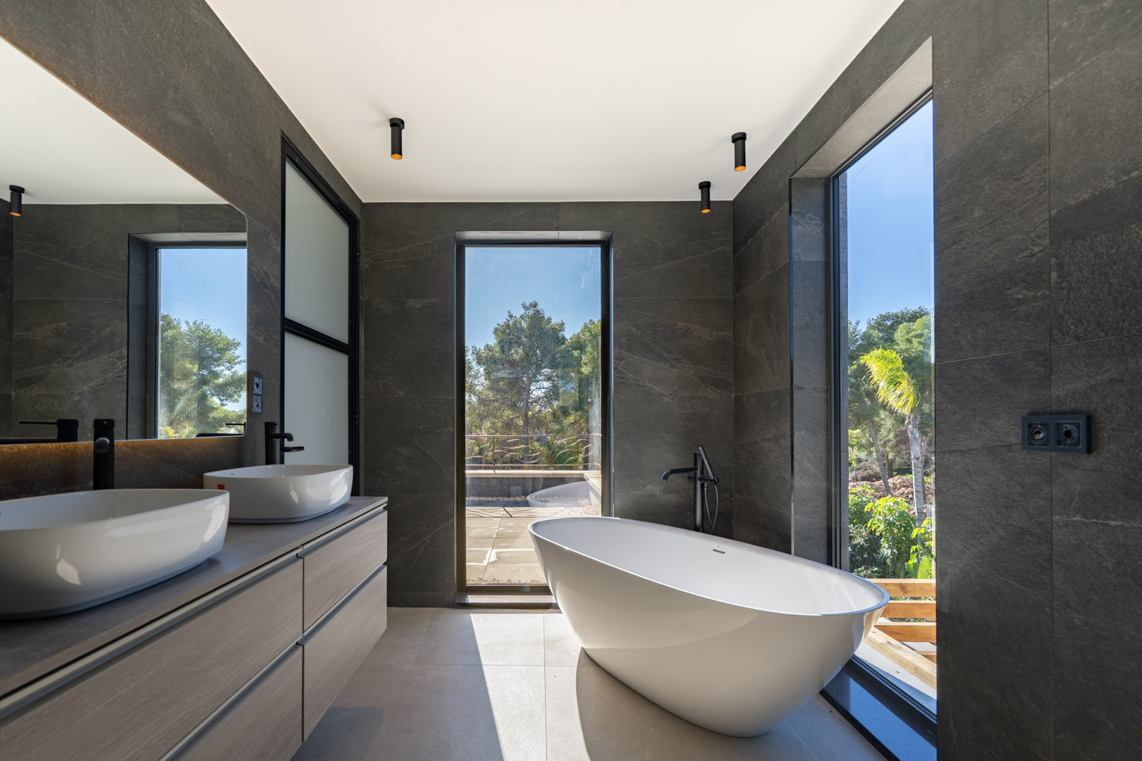 Baño principal con bañera exenta, lavabo doble sobre mueble de madera y grandes ventanales con vistas al jardín. Destacan los revestimientos en piedra oscura y la grifería negra.