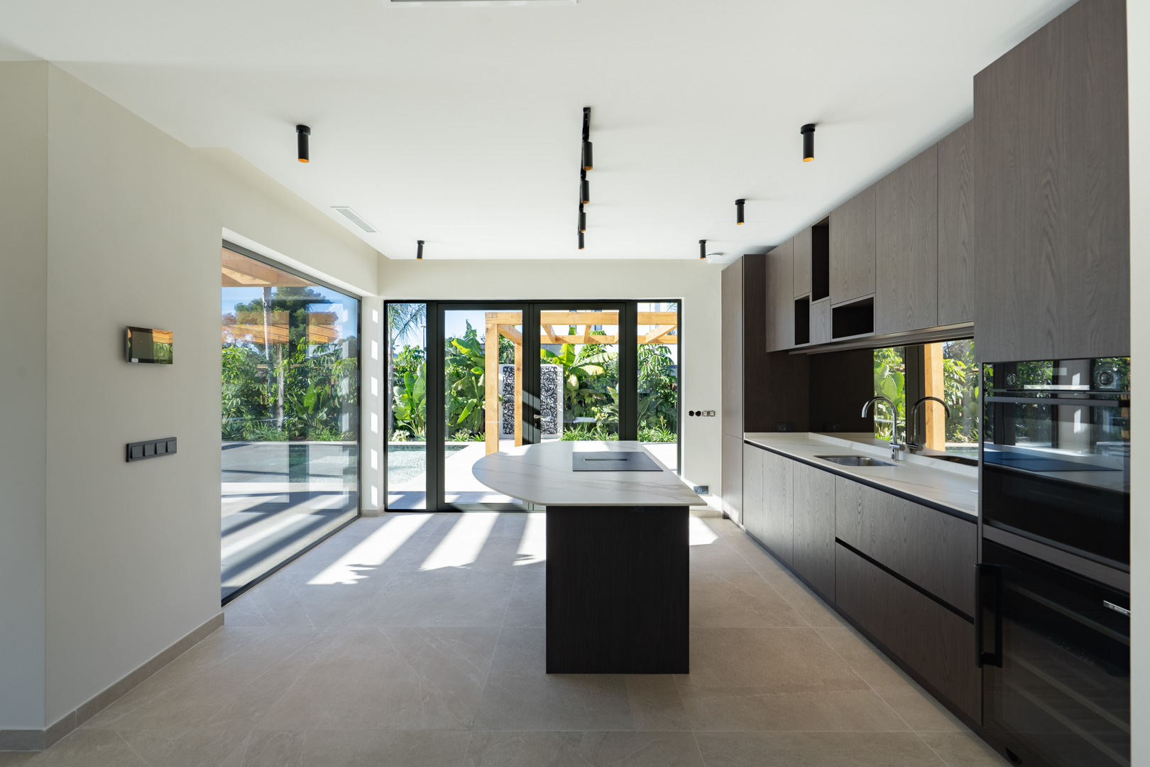 Cocina moderna con isla central, encimeras de piedra blanca y armarios de madera oscura. Grandes ventanales conectan el espacio con el jardín y la piscina exterior.