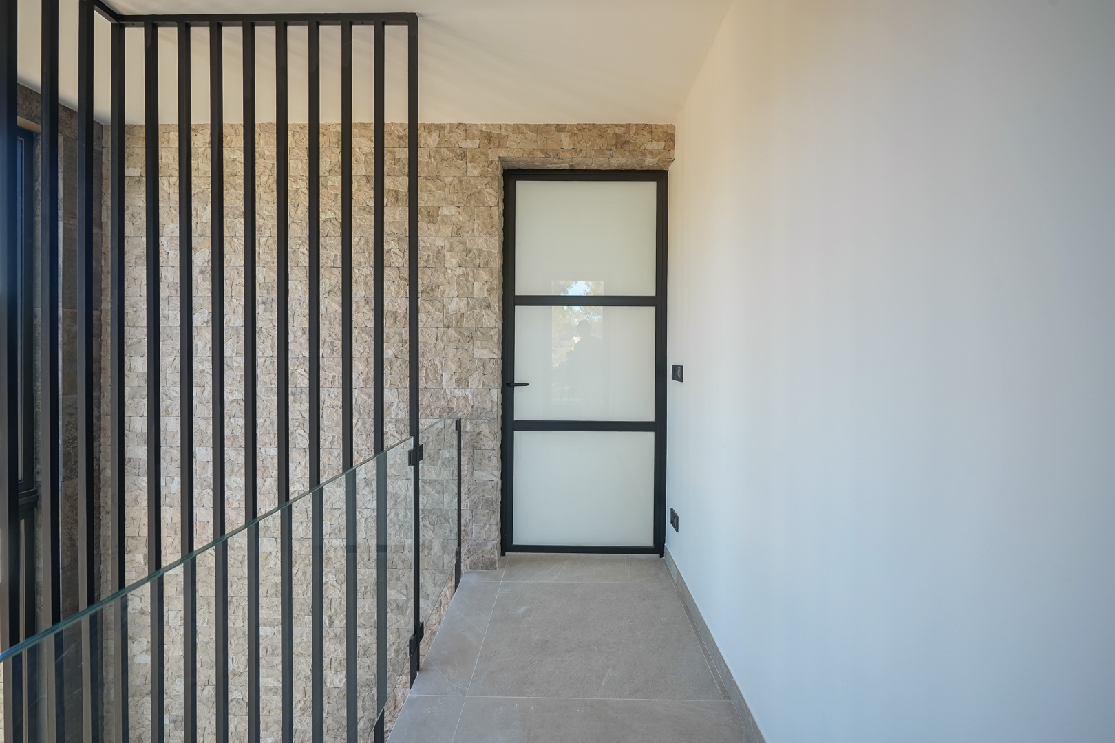 Distribuidor de la planta superior con pared de piedra natural, barandilla de cristal con listones negros y puerta de estilo industrial con cristal traslúcido.