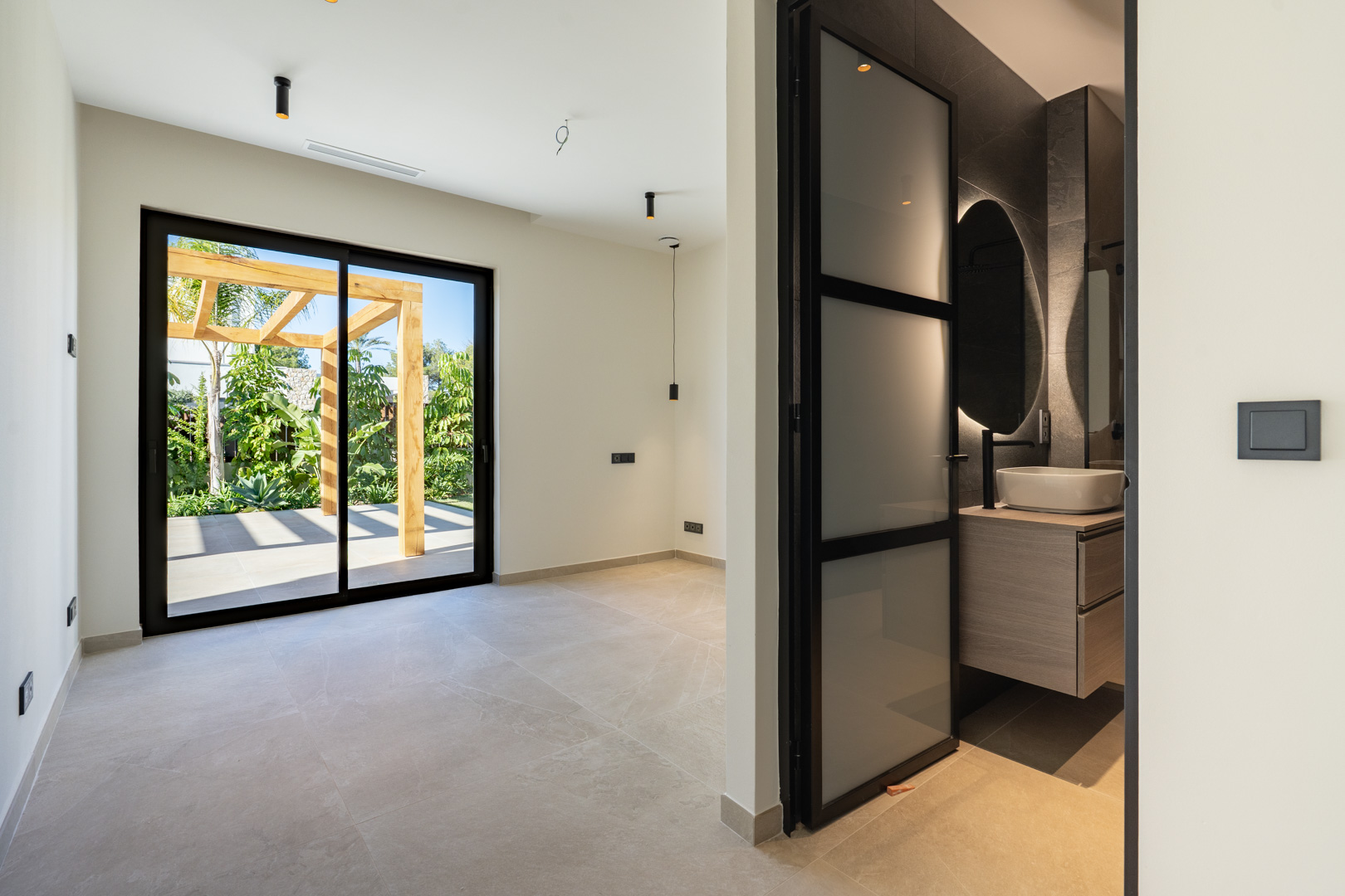 Dormitorio con acceso a terraza y pérgola de madera. Incluye baño en suite con lavabo sobre encimera, espejo retroiluminado y carpintería de aluminio negro.