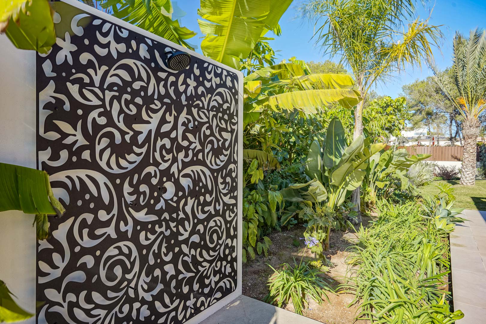 Ducha exterior con panel decorativo troquelado en negro, integrada en un jardín mediterráneo con palmeras y plantas tropicales.