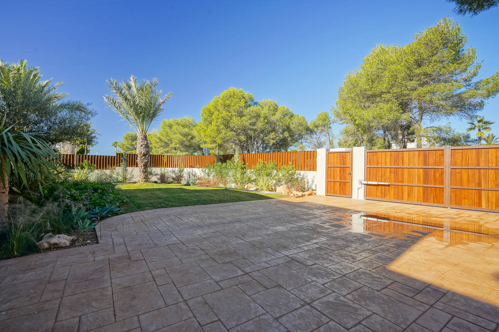 Entrada principal con portón automático de madera, zona de aparcamiento pavimentada y jardín mediterráneo diseñado con palmeras y césped natural.
