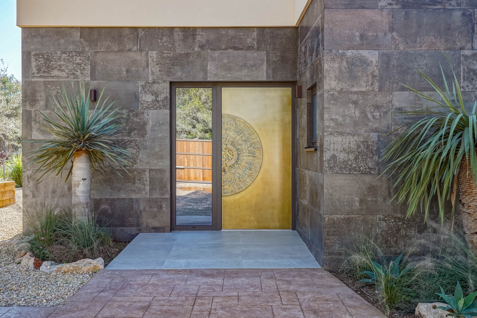 Entrada principal con puerta de diseño en tonos dorados y relieve circular, enmarcada por muros de piedra oscura y vegetación mediterránea.