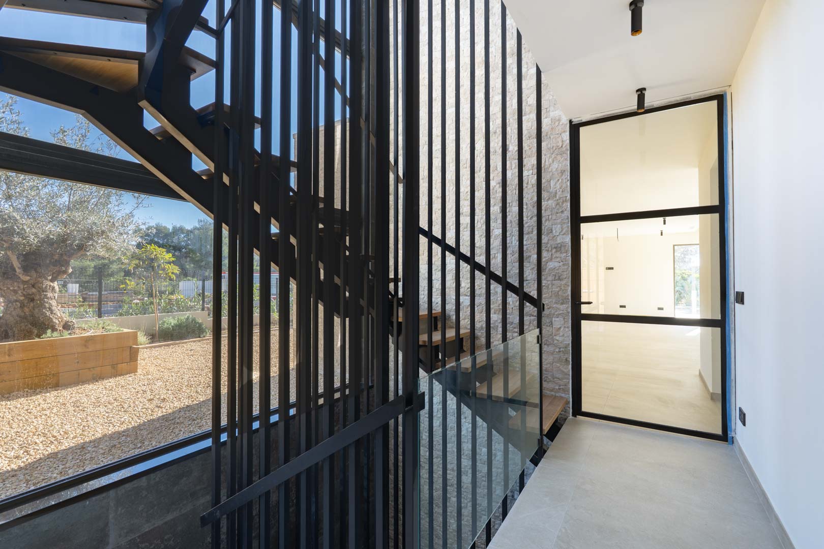 Escalera de diseño con peldaños de madera y barandilla de listones metálicos negros, junto a un gran ventanal con vistas al jardín mediterráneo y muro de piedra natural.