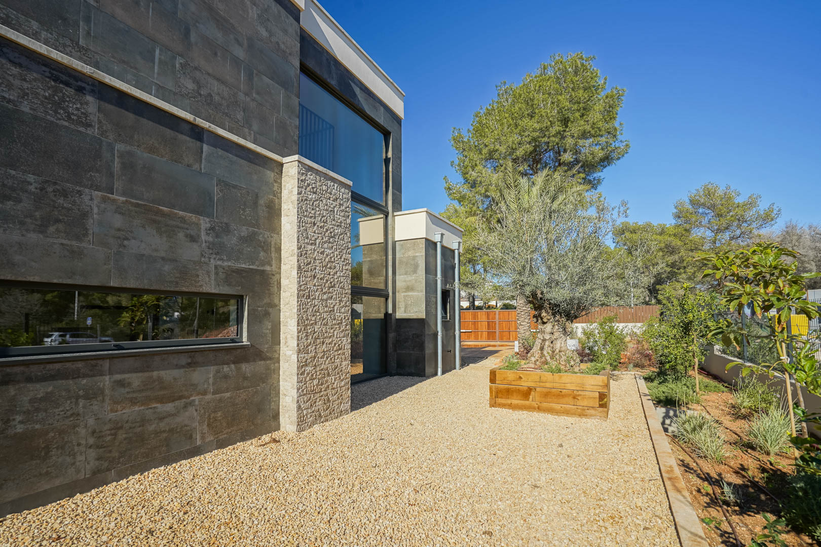 Fachada moderna con revestimiento de piedra oscura y pilares de piedra natural, grandes ventanales y jardín mediterráneo con olivo centenario en jardinera de madera.