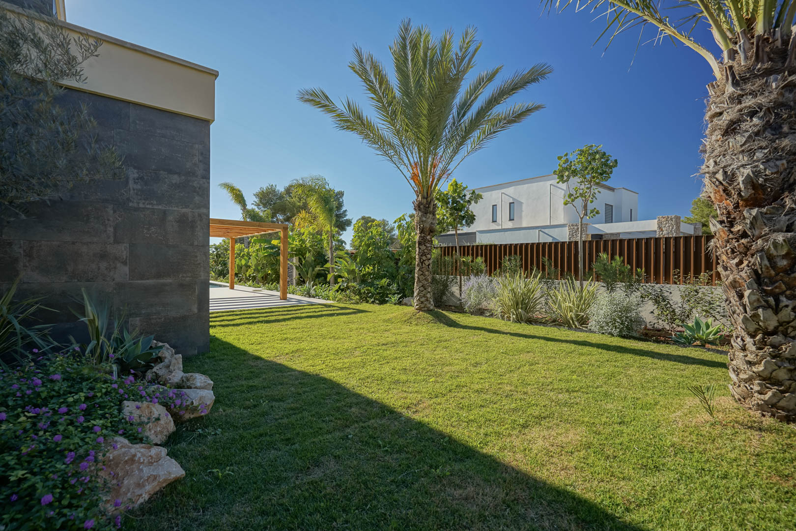 Jardín mediterráneo con césped natural, palmeras y vegetación autóctona junto a una fachada de piedra oscura y una pérgola de madera de iroko.