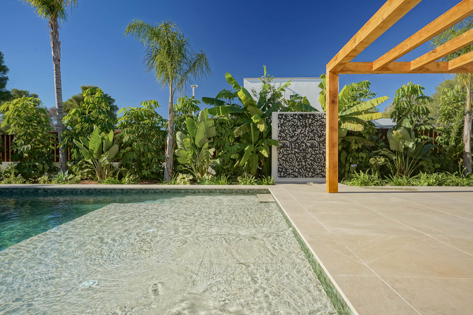 Piscina de agua salada estilo Bali con zona de playa, rodeada de una terraza de gres porcelánico, pérgola de madera de iroko y un exuberante jardín tropical.