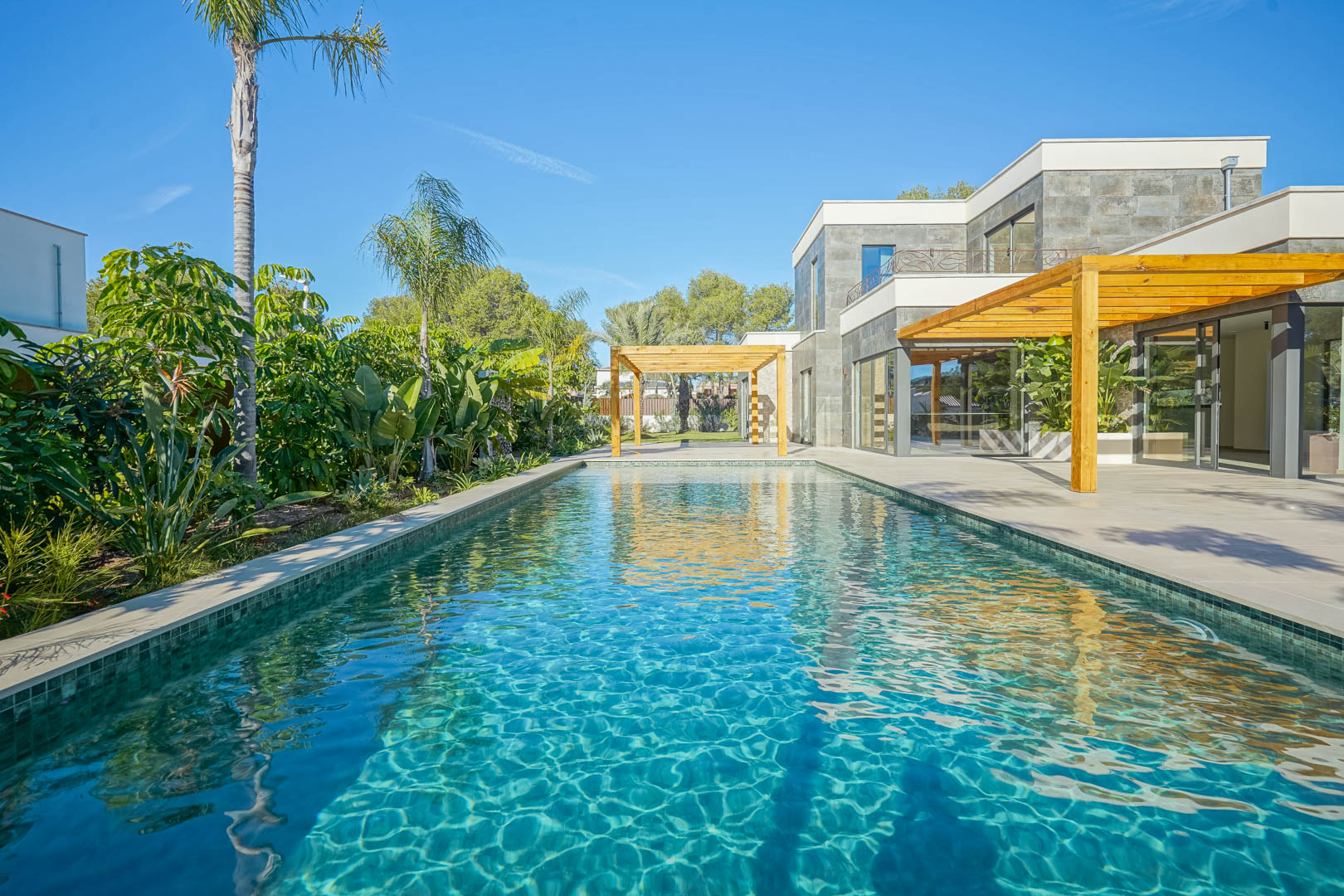 Piscina de estilo Bali con azulejos verdosos, rodeada de una amplia terraza con pérgolas de madera de iroko y un jardín mediterráneo maduro.