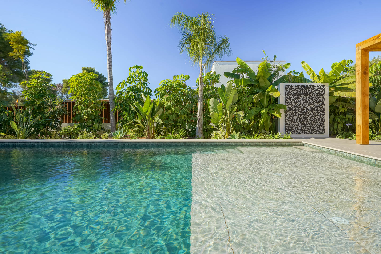 Piscina de estilo Bali con zona de playa y gresite turquesa, rodeada de un jardín mediterráneo exuberante con palmeras y paneles decorativos.
