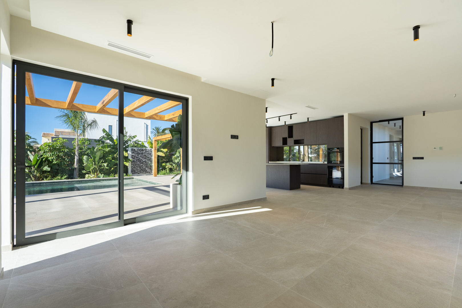 Salón diáfano con suelos porcelánicos y grandes ventanales que conectan con la terraza, la pérgola de iroko y la piscina. Al fondo, cocina de diseño en madera oscura.