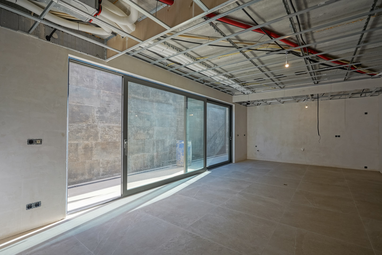 Sótano diáfano en construcción con grandes ventanales correderos, suelo porcelánico de gran formato y preinstalaciones de climatización y electricidad visibles en el techo.
