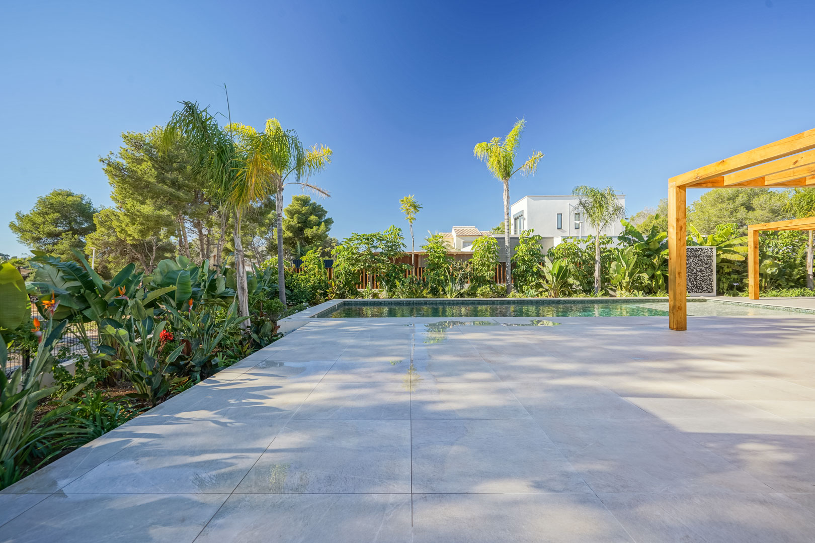 Terraza de gres porcelánico junto a una piscina estilo Bali, rodeada de un jardín mediterráneo con palmeras y pérgolas de madera de iroko.