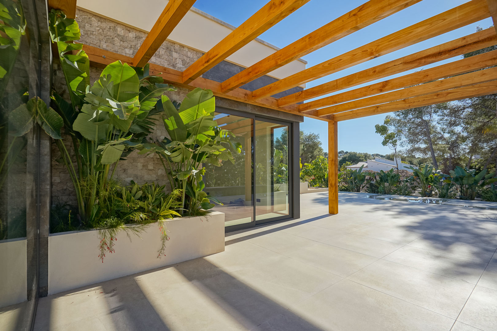 Terraza exterior con pérgola de madera de iroko, suelos de gres porcelánico y jardinera integrada con plantas tropicales junto a la fachada de piedra.