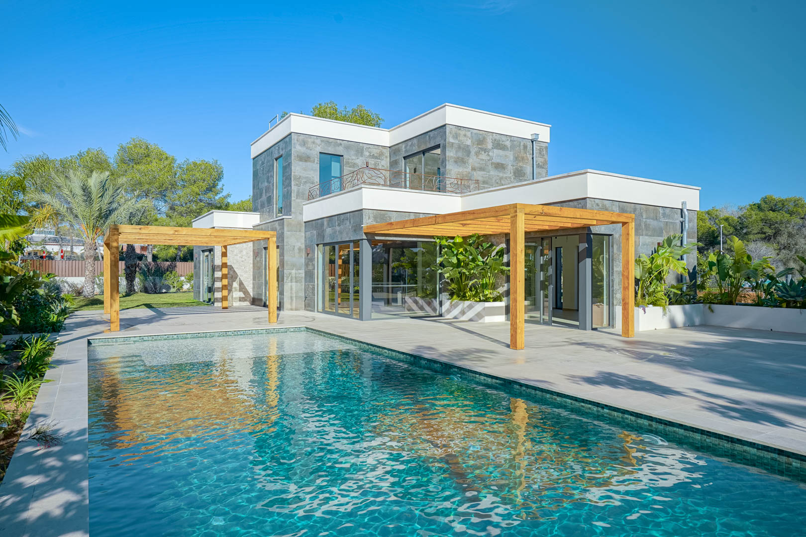 Vista exterior de la villa con piscina de estilo Bali, amplias terrazas y pérgolas de madera de iroko integradas en una fachada con revestimiento de piedra natural.