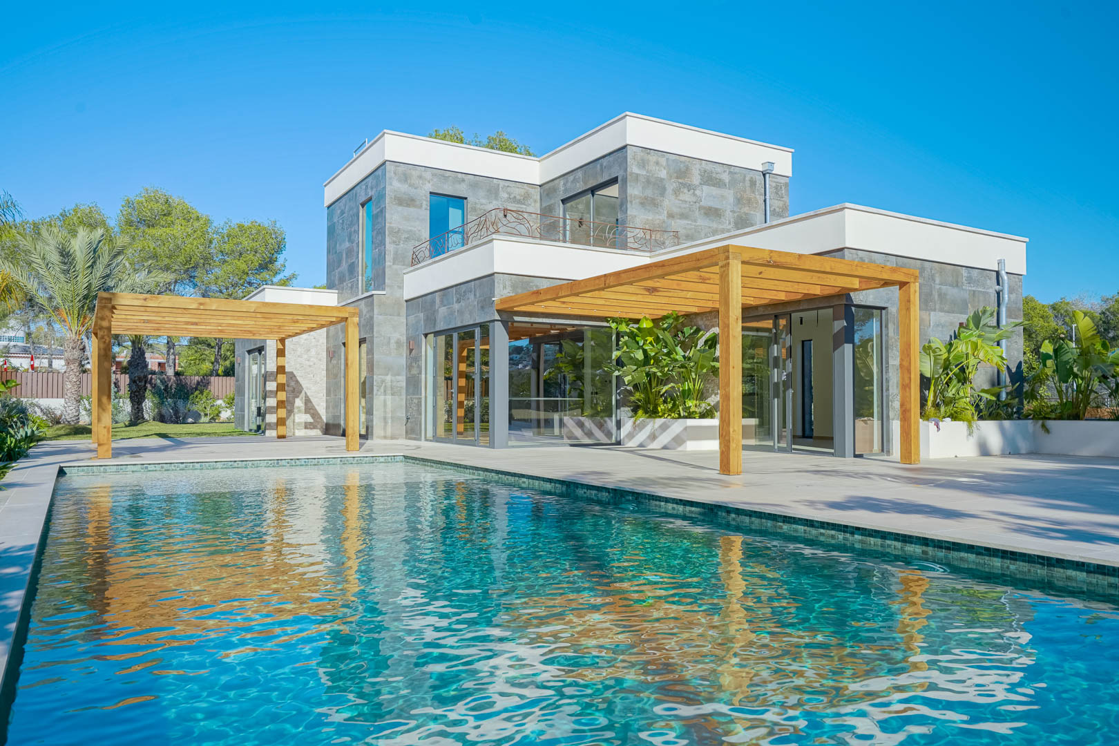 Vista exterior de la villa con piscina de mosaico turquesa, pérgolas de madera de iroko y fachada de piedra natural con amplios ventanales.