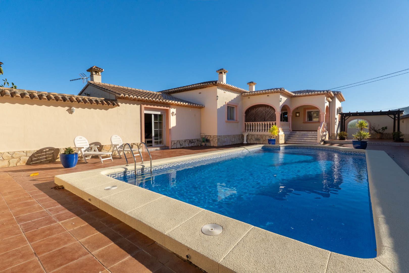 Amplia zona exterior con piscina privada, rodeada de una terraza de gres y una naya con arcos de estilo mediterráneo bajo un cielo despejado.