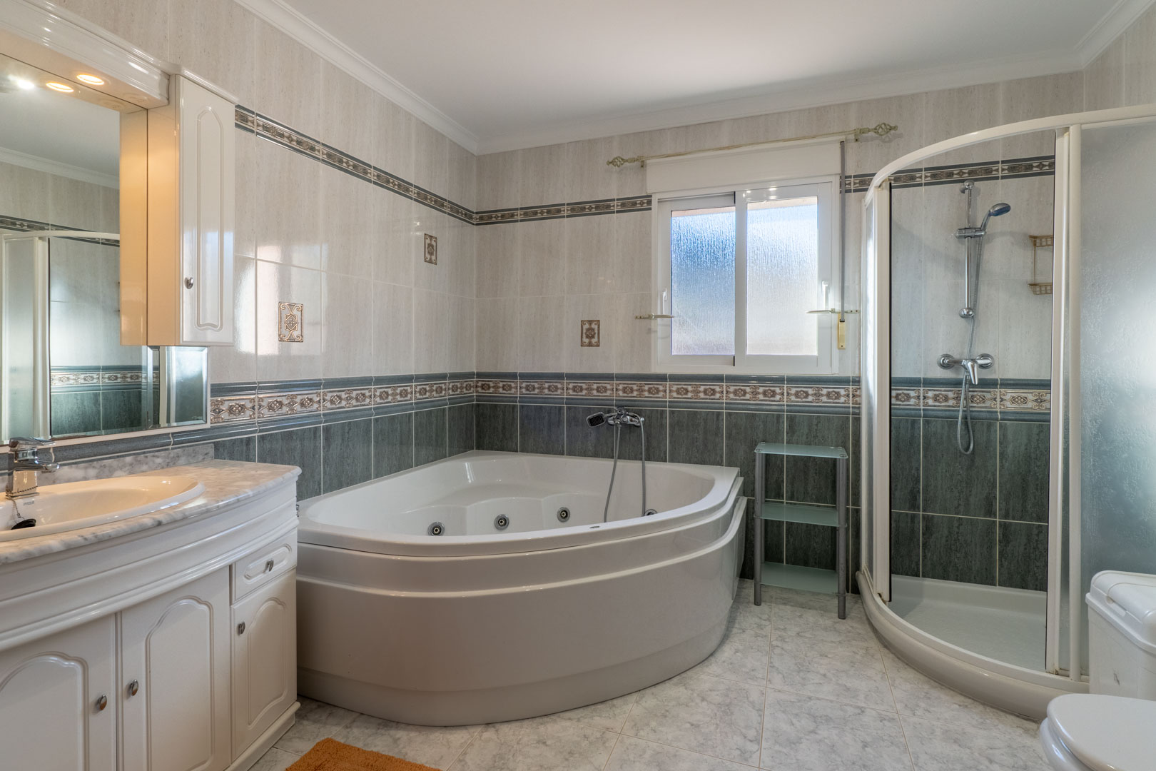 Baño completo con bañera de hidromasaje rinconera, ducha independiente con mampara curva y mueble de lavabo con encimera de mármol y azulejos decorativos.