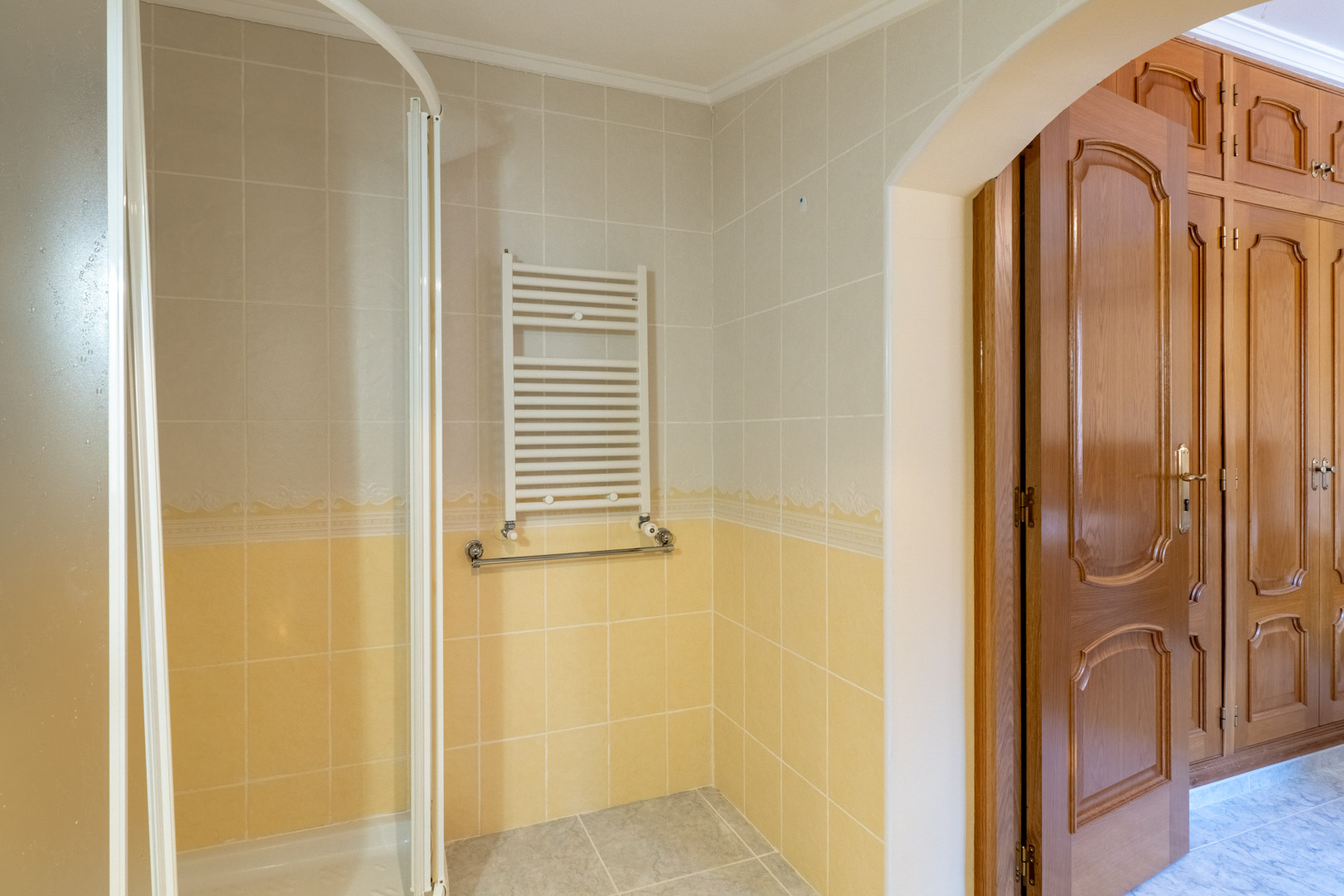 Baño con azulejos bitonales, radiador toallero y ducha, conectado mediante un arco al dormitorio con amplios armarios empotrados de madera tallada.