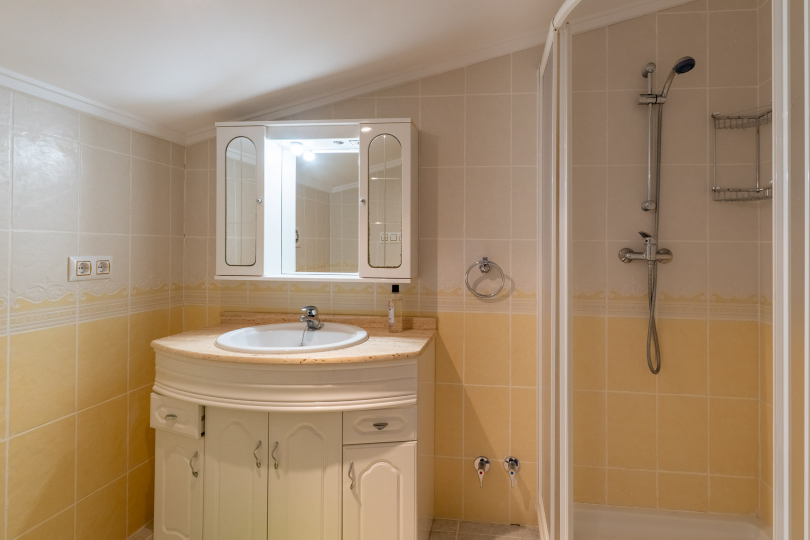 Baño funcional con lavabo encastrado en encimera de mármol, mueble de almacenamiento blanco y zona de ducha con mampara, revestido en azulejos de tonos crema.