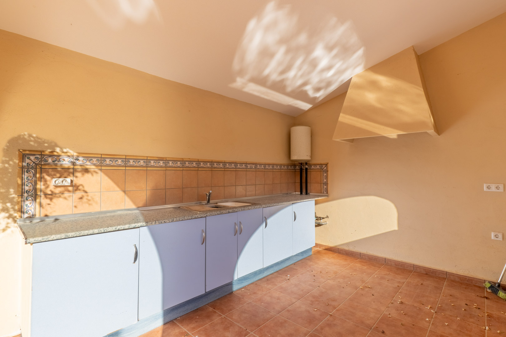 Cocina exterior funcional con encimera de granito, armarios inferiores en azul claro y frente de azulejos tradicionales. Incluye fregadero de acero y campana extractora integrada.