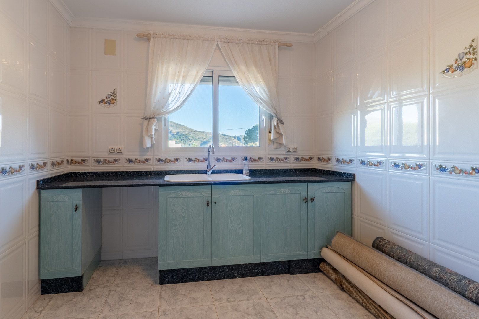 Cocina funcional con armarios de madera en tono azul pastel, encimera de granito oscuro y ventana con vistas despejadas a las montañas de Jalón.