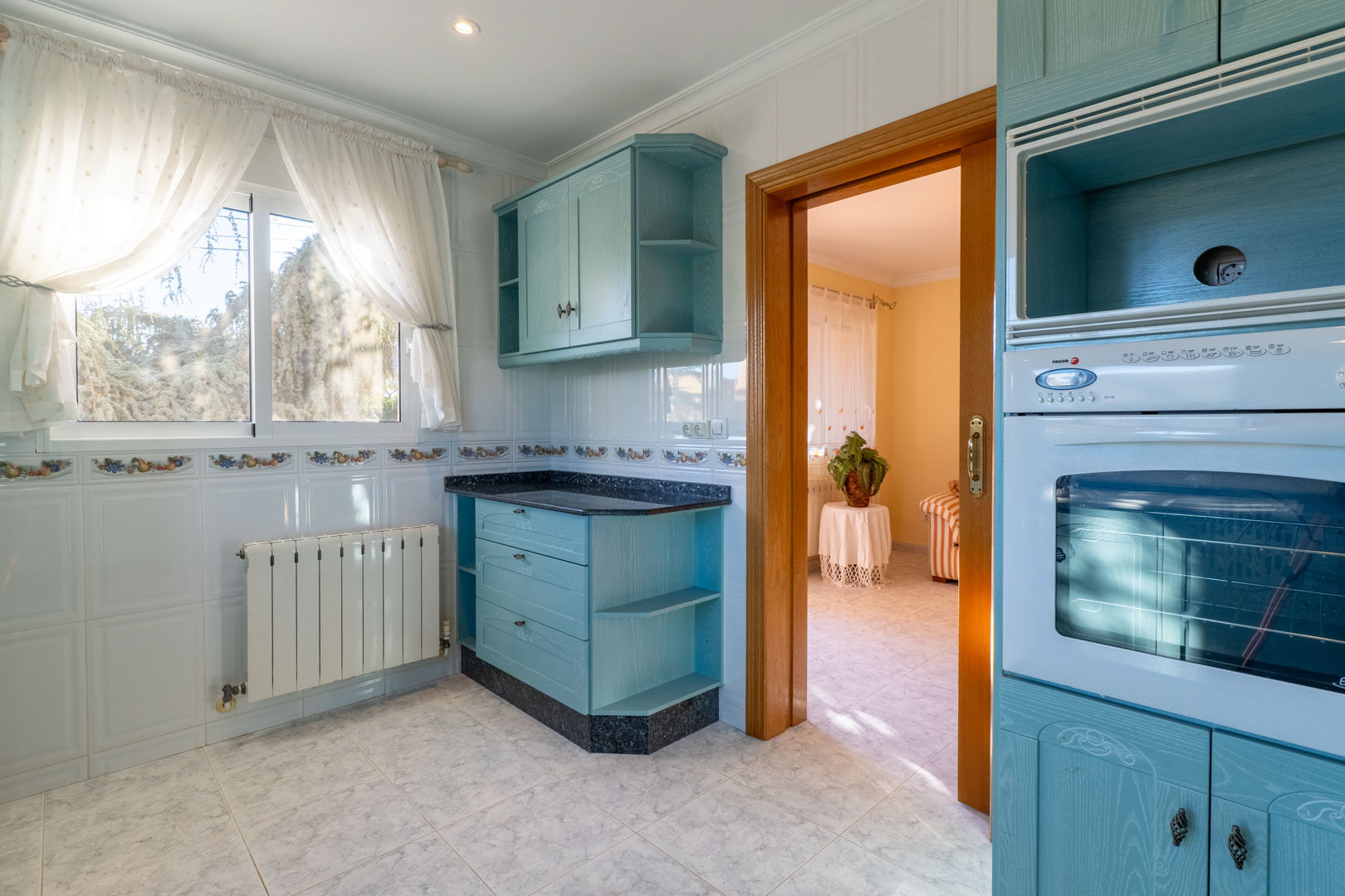 Cocina independiente con mobiliario en azul suave, encimera de granito oscuro, paredes alicatadas en blanco y acceso directo al salón a través de un marco de madera.